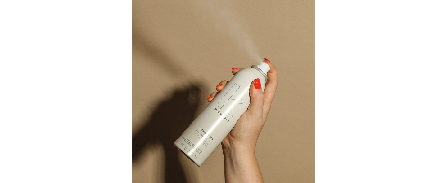 A hand with red nail polish holding a white aerosol can of Kevin Murphy FRESH.HAIR dry shampoo spray, against a beige background.